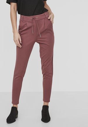 Pantalon pour femme bordeaux, ajusté, avec une taille à cordon, des plis à l'avant et des poches latérales, fabriqué à partir d'un tissu extensible. Bottines noires aux chevilles portées.