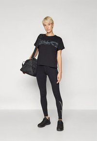 Melns grafisks T-krekls ar zilām līnijām, melnas legingi ar Fakturētu balto dizainu un melnas sporta kurpes. Pār plecu ir uzlikts melns somiņa.