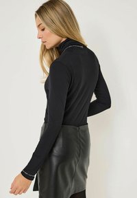 Blondýnka s dlouhými vlasy má na sobě černý rolák a černou koženou sukni, stojí bokem na jednoduchém světlém pozadí.