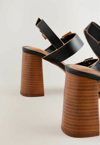 Sandali con tacco in pelle nera, con fasce larghe e accessori in oro. Presentano un tacco conico marrone scanalato e una soletta color sabbia.