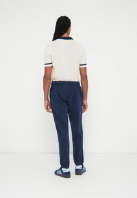 Camisa polo beige con cuello y rayas en las mangas en navy, combinada con pantalones de chándal navy. Modelo lleva zapatillas azules con calcetines blancos.