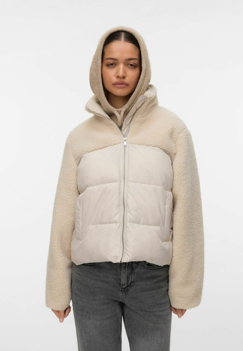 Kvinde iført beige sherpa- og quiltet jakke med hættetrøje under, kombineret med mørkegrå jeans, stående foran en ensfarvet baggrund.