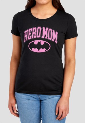 Woman wearing black t-shirt with pink "HERO MOM" text and pink Batman logo, paired with light blue jeans on gray background.