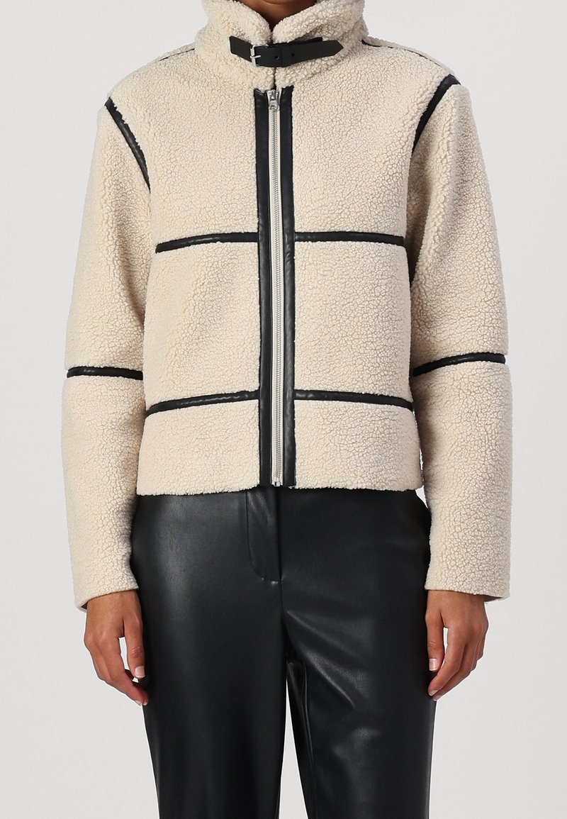 Veste sherpa beige avec bordures noires et fermeture éclair, portée avec un pantalon en cuir noir, visible du cou jusqu'aux hanches, les mains le long du corps.