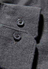 Donkergrijze stof met een getextureerde weving, voorzien van twee zwarte knopen en een puntige kraag. Close-up toont details van de stiksels en knoopsgaten.