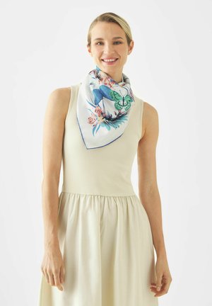 Femme souriante portant une robe crème sans manches et une écharpe blanche avec des imprimés papillons bleus, verts et floraux autour du cou.