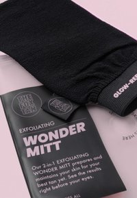 Gant exfoliant noir en tissu texturé, avec un poignet élastique et une étiquette. Accompagné d'un emballage en carton noir avec un texte rose.