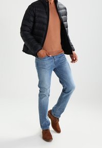 Giacca a vento nera, maglione marrone, jeans azzurri chiari e scarpe marroni. La giacca ha una trapuntatura orizzontale, il maglione ha il collo a costine.