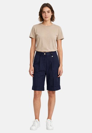 Donna in piedi che indossa una maglietta beige a maniche corte, pantaloncini blu scuro al ginocchio con dettagli a bottoni e scarpe da ginnastica bianche su sfondo bianco.