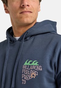 Marine hoodie met een voorzijdegrafiek van groene golven en de tekst "BILLABONG FEELING FREE 73" in roze en oranje. Gemaakt van een katoenmix.