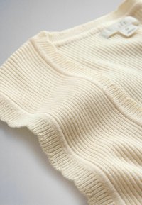 Top de tirantes de punto acanalado color crema con escote cuadrado y bordes ondulados, colocado sobre una superficie blanca.