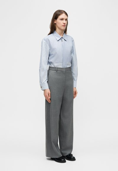 Femme debout portant une chemise à rayures bleues et blanches boutonnée, rentrée dans un pantalon gris à taille haute et jambes larges, avec des chaussures noires.