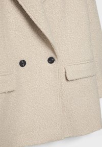 Beige textuur tweedelige jas met zwarte knopen, voorzien van twee voorkruiszakken en een brede reversontwerp. Zacht, fluffy materiaal.