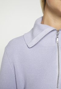 Maglione zip-up di colore viola chiaro realizzato in tessuto a coste, con un ampio colletto e dettagli in zip argentata.