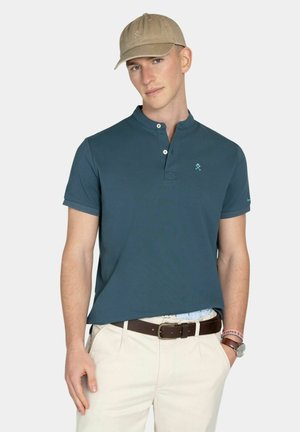 Giovane uomo che indossa una polo a maniche corte color verde acqua con due bottoni, cappello beige, pantaloni beige chiaro, cintura marrone, orologio e braccialetti, in piedi contro uno sfondo bianco.
