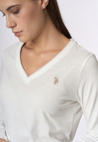 T-shirt bianco a maniche lunghe con scollo a V e logo ricamato in oro sul lato sinistro del petto. Tessuto morbido e design semplice.