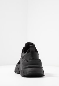 Baskets noires avec une tige texturée en cuir et mesh, dotées d'une semelle épaisse, d'un talon renforcé et d'une languette à l'arrière.