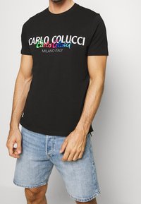 Camiseta de algodón negra con texto de logo multicolor "CARLO COLUCCI" y "MILANO ITALY". Combinada con pantalones cortos de denim claros, mostrando un diseño ajustado.