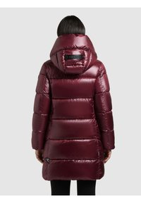 Cilvēks, valkājot garu, spīdīgu burgundijas krāsas pūkainu mēteli ar lielu kapuci, skatoties no aizmugures pret vienkāršu gaišu fona.