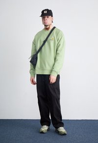 Grøn sweatshirt med logo, sort crossbody taske, sorte bukser og grågrønne sneakers. Glat stof og afslappet pasform.