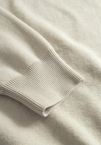 Détail d'un pull beige en maille avec un bord-côte à la manche, une texture douce et un tissu lisse présentant de légères variations dans le tissage.