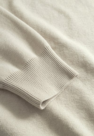 Détail d'un pull beige en maille avec un bord-côte à la manche, une texture douce et un tissu lisse présentant de légères variations dans le tissage.