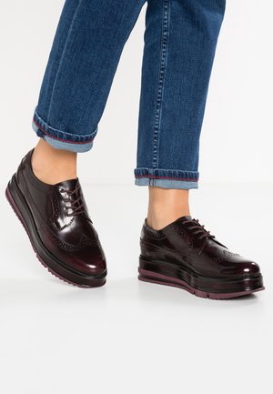 Persona que lleva zapatos de plataforma brogue de cuero burdeos oscuro con suelas gruesas y vaqueros azules arremangados sobre un suelo blanco.