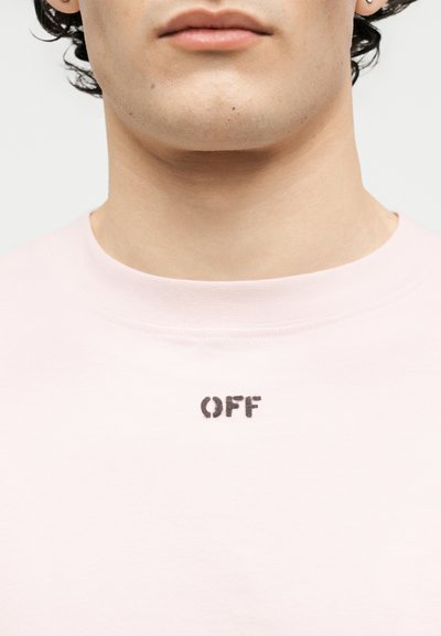 Κοντινό πλάνο του κάτω μέρους του προσώπου και του λαιμού ενός ατόμου που φοράει ροζ ανοιχτό πουκάμισο με τη λέξη «OFF» τυπωμένη με μαύρα γράμματα στην περιοχή του γιακά.