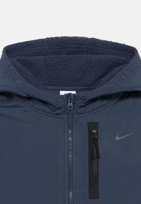 Casaco azul marinho com capuz forrado a fleece, fecho frontal e bolso lateral com acessórios em preto contrastante. Textura suave, design desportivo.