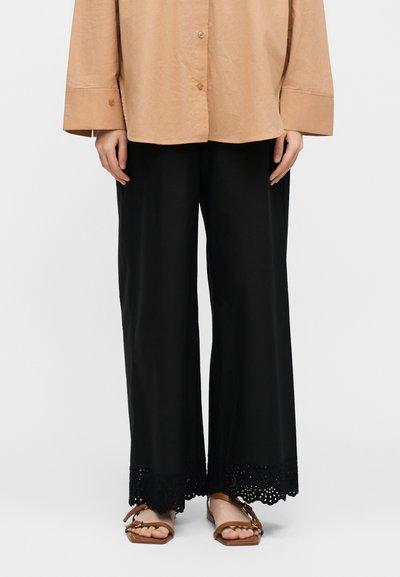 Άτομο στέκεται φορώντας φαρδιά μαύρα παντελόνια με δαντελένιο τελείωμα, μπεζ πουκάμισο με κουμπιά και καφέ σανδάλια με λουράκια και αγκράφες.