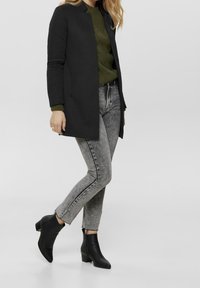 Manteau long noir porté sur un pull vert texturé, associé à un jean skinny gris et des bottines noires. Tissu lisse et coupe ajustée.