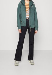 Green, hooded jacket with zippers and contrasting stitching, paired with a striped shirt, dark flared jeans, and gray sneakers.