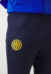 Pantaloni sportivi blu navy con vita elastica; presentano un logo ricamato giallo circolare sulla coscia. Tessuto liscio e leggero.