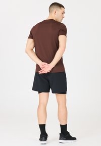 Ruskea istuva lyhythihainen paita yhdistettynä mustiin juoksushortseihin. Malli on selin, mikä tuo esiin yksinkertaisen ja suoran siluetin.