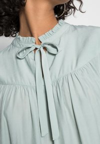 Blouse vert menthe clair en tissu doux, présentant un col froncé, une fermeture à nœud et des détails froncés sous la poitrine.