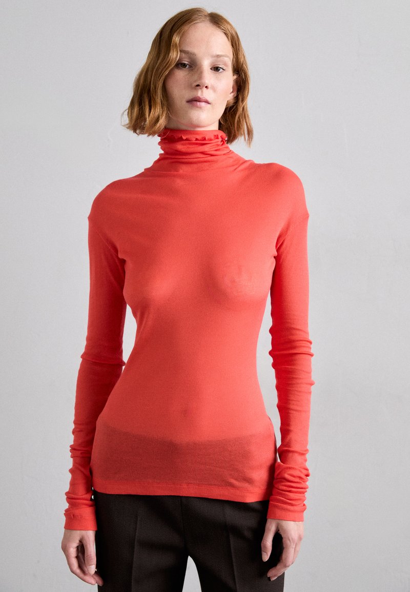 Red ribbed turtleneck sweater with long sleeves and a ruffled collar. Fabric appears stretchy and fitted, paired with dark brown trousers.
