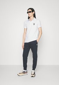Hvit polo skjorte med svart logo, mørke joggebukser med logo, og beige-svarte sneakers; modell iført mørke solbriller.