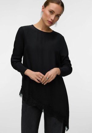 Femme à la peau claire et aux cheveux relevés porte un long haut noir asymétrique avec bordure en dentelle et un pantalon foncé, tenant les mains devant elle.