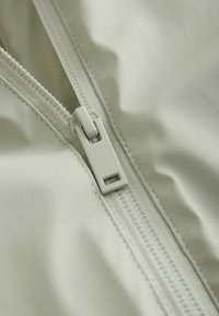 Gaiši pelēks audums ar gludu tekstūru, kas rotāts ar rāvējslēdzēju un šūšanas detaļām, tuvplāns rāvējslēdzēja vilkšanas cilnei un apkārtējam materiālam.