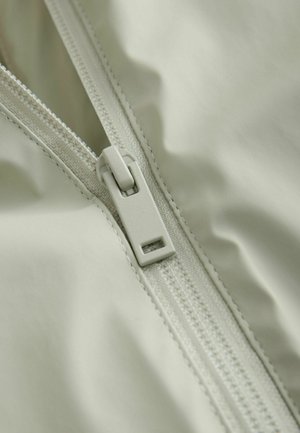 Material textil gri deschis cu o textură netedă, prevăzut cu un fermoar și detalii de cusătură, prim-plan al cursorului fermoarului și materialului înconjurător.