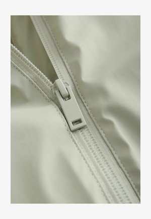 Világosszürke, sima tapintású szövet, cipzárral és varrási részlettel, közelről a cipzár húzójára és a környező anyagra fókuszálva.