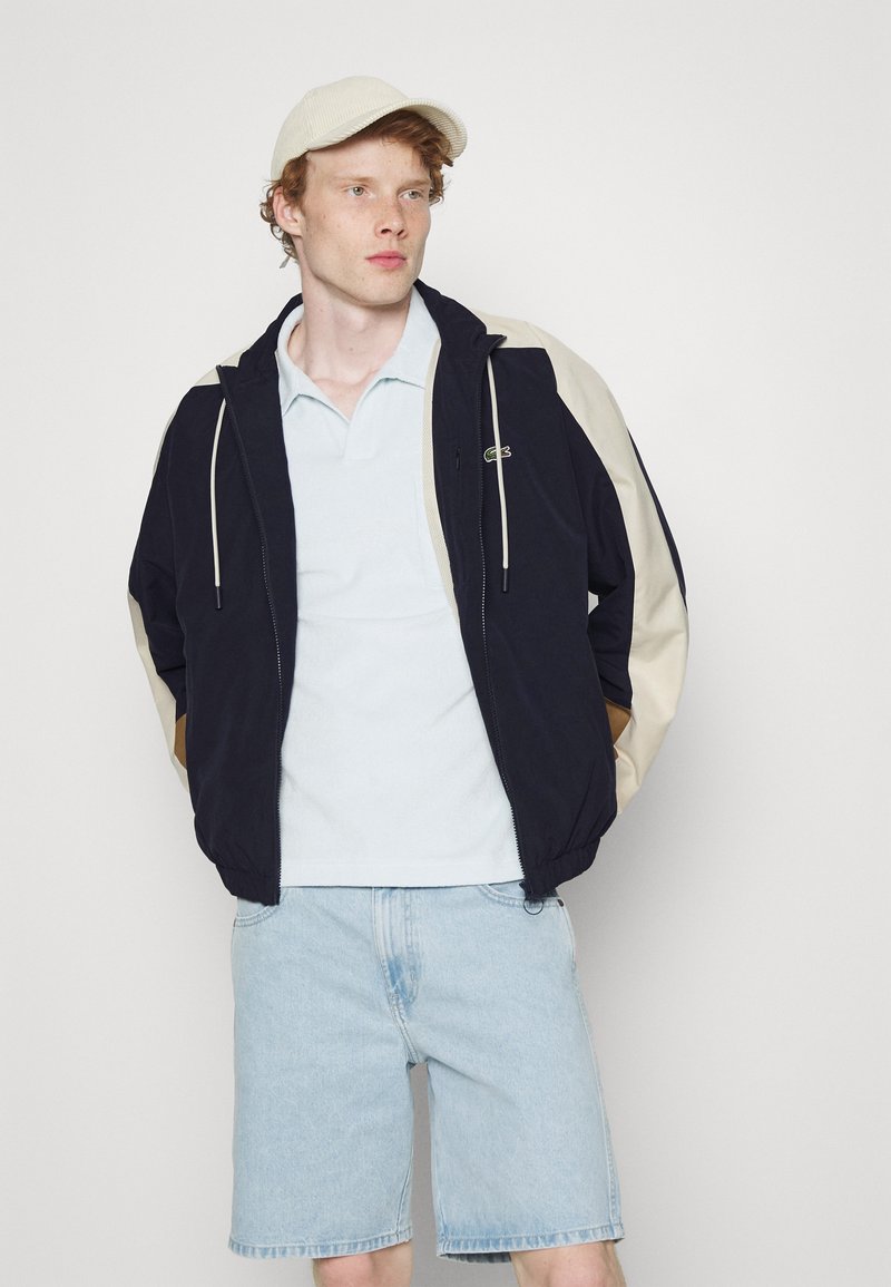 Marineblauwe en crème kleurgeblokte windbreaker met verstelbare trekkoorden, ritssluiting aan de voorkant en logo-accent. Lichtblauwe polo en denim short gedragen.