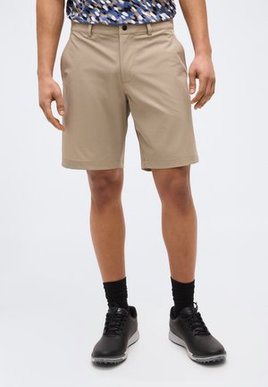 Mężczyzna ubrany w beżowe, sięgające do kolan krótkie spodenki, czarne skarpetki i czarne tenisówki, stoi na tle jasnego, jednolitego tła.