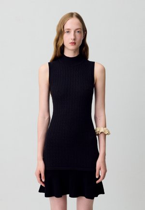 Abito in maglia nera caratterizzato da un collo alto, texture a coste e silhouette aderente, con orlo svasato e un accento di tessuto chiaro sul polso.