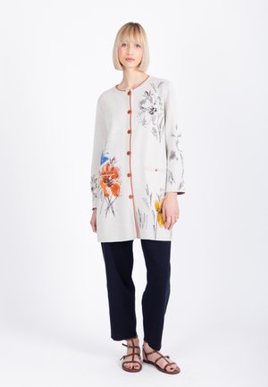 Kvinde iført en lang cremefarvet cardigan med blomsterprint, brune knapper, sorte bukser og brune remsandaler, stående på en hvid baggrund.