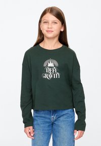 Zöld hosszú ujjú, pamutból készült crop top fehér "DEFY GRAVITY" felirattal, melyen egy kastély és napsugár motívum található. Kék farmerral párosítva.