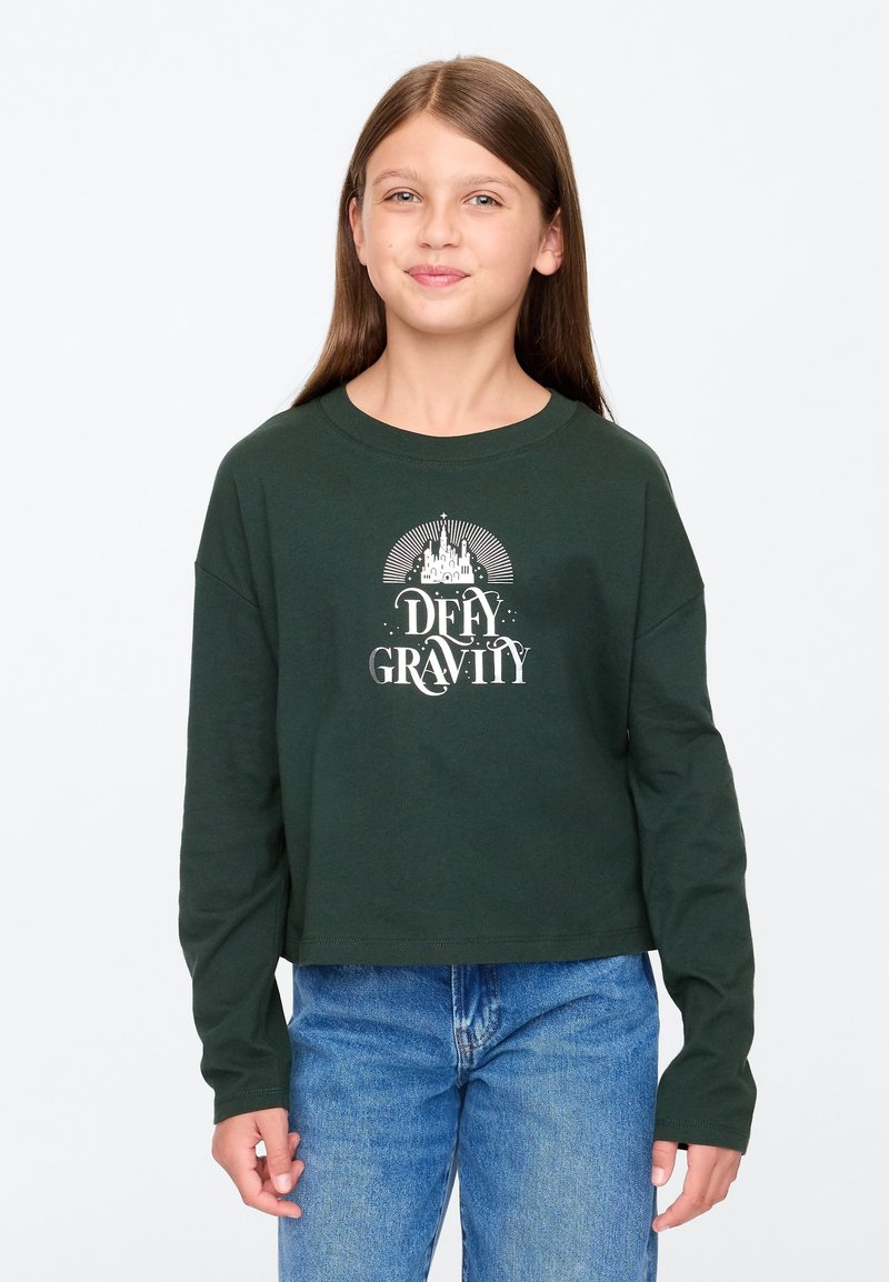 Zöld hosszú ujjú, pamutból készült crop top fehér "DEFY GRAVITY" felirattal, melyen egy kastély és napsugár motívum található. Kék farmerral párosítva.