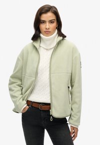 Giacca in pile verde chiaro con colletto alto, zip frontale e tasche laterali, indossata sopra un maglione bianco a trecce e jeans scuri.