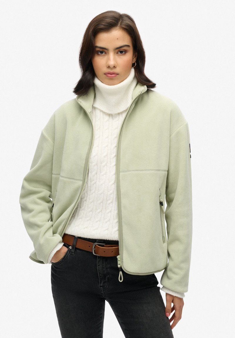 Giacca in pile verde chiaro con colletto alto, zip frontale e tasche laterali, indossata sopra un maglione bianco a trecce e jeans scuri.