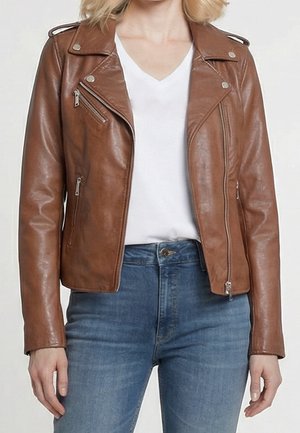 Femme portant une veste en cuir marron par-dessus un t-shirt blanc à col en V et un jean bleu, montrée des épaules aux hanches sur un fond uni.